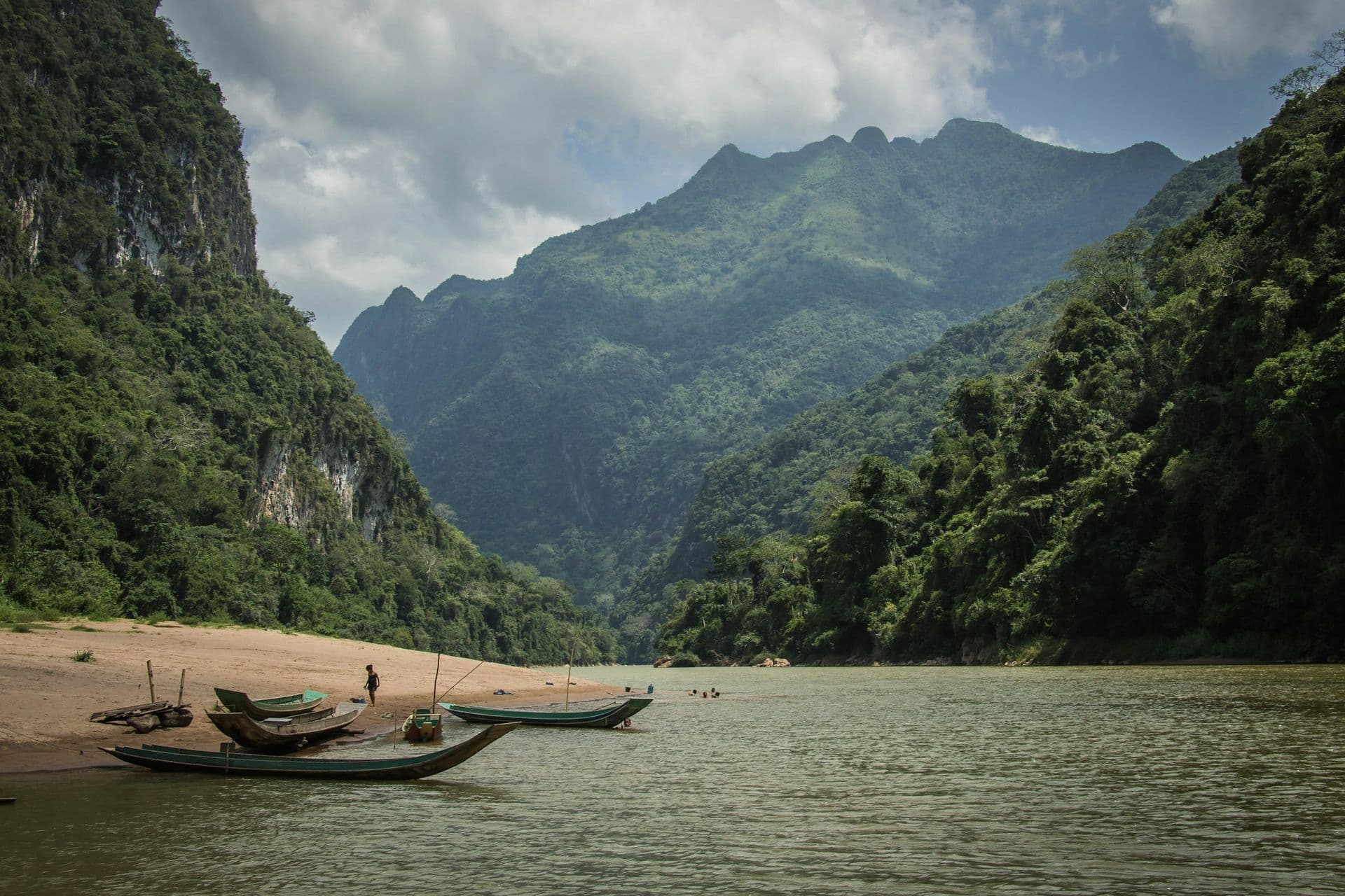 Wooden longboats pulled up on a river beach beneath steep forested mountains