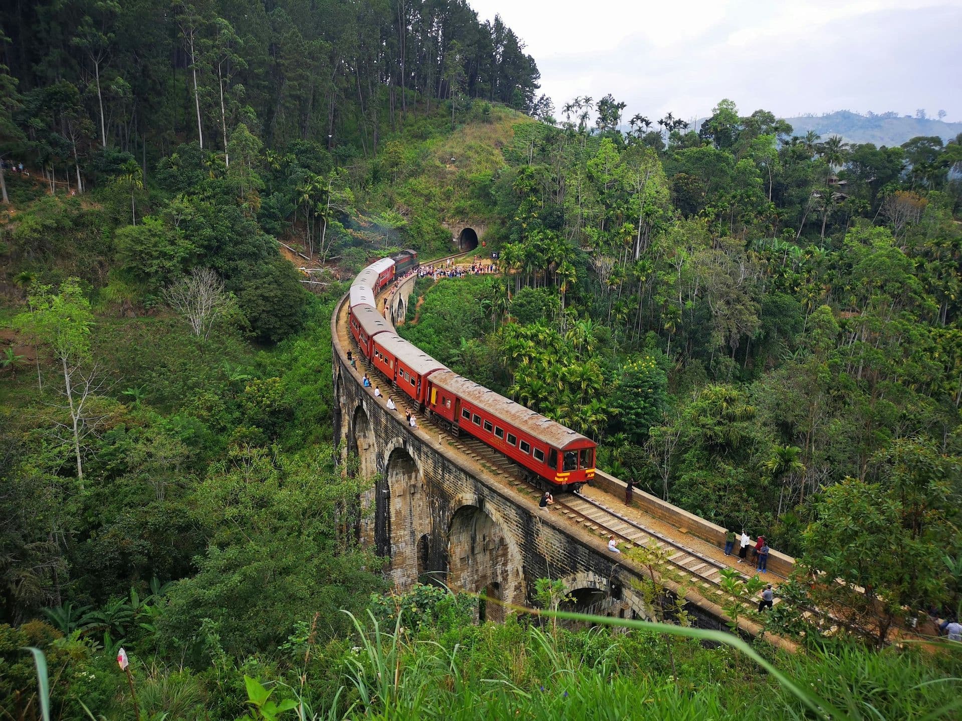 A red train crossing the stone arches of the Nine Arch Bridge through green jungle near Ella